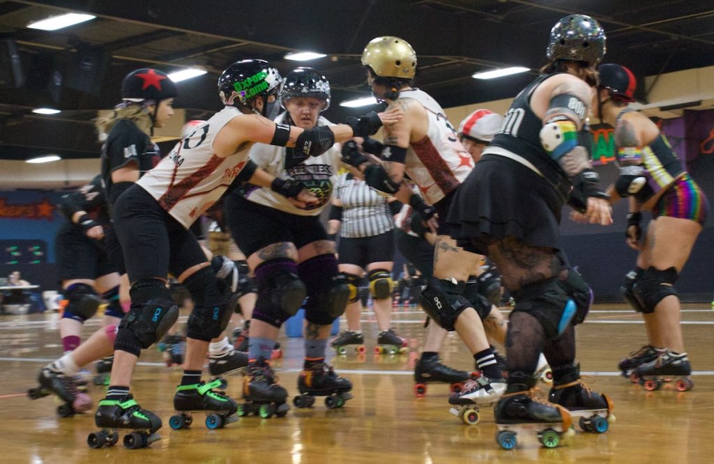 Skates and shoulders collide at Wheels during a fast-paced jam, while teammates work to clear a path for their jammer.