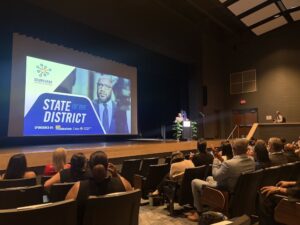 People in an auditorium listen to a speech delivered from the stage. A large screen reads "State of the District."