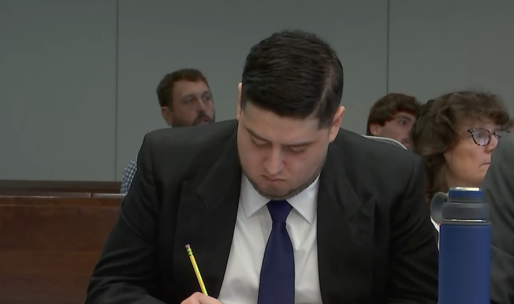 A man in a black blazer sits in court, writing with a pencil.